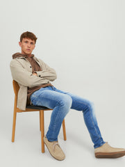 رجل يجلس على كرسي مرتديًا بنطلون جينز سلم ستريت فيت أزرق، مع سترة بيج وتيشيرت بني وحذاء كريمي.