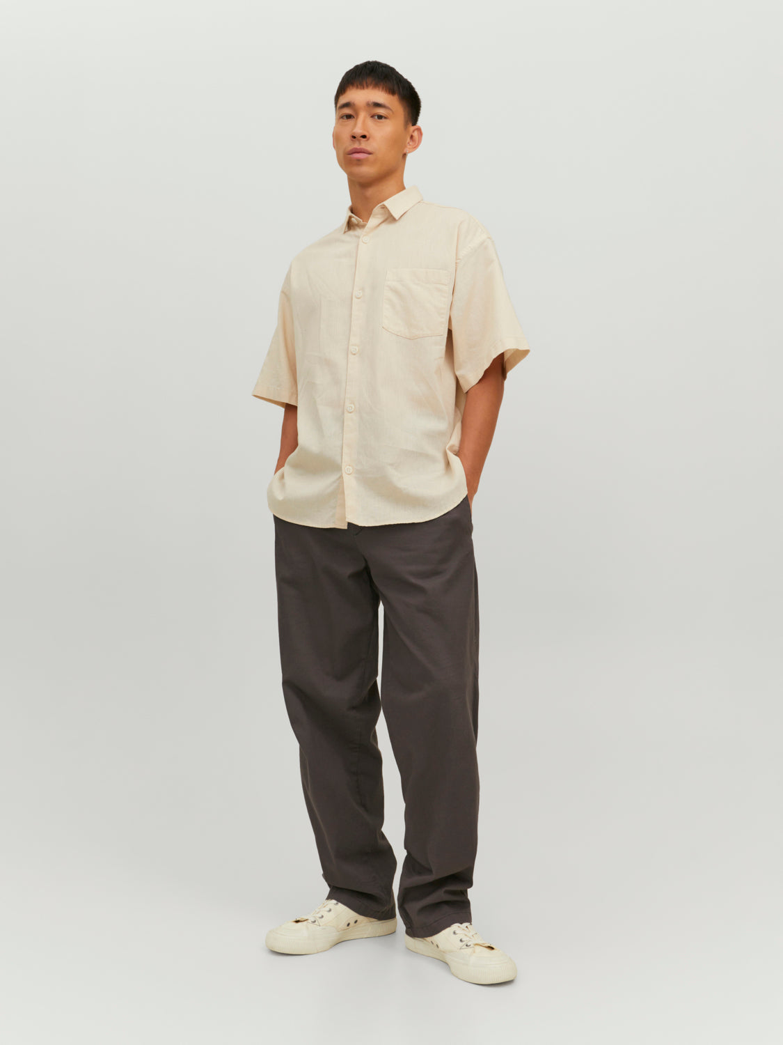 رجل يرتدي قميص نص كم بيج فاتح وبنطال رمادي، واقف بكامل طوله أمام خلفية بيضاء