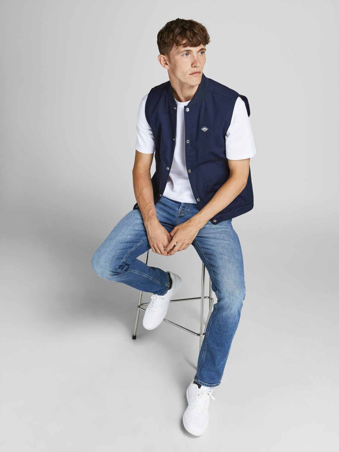 رجل يرتدي بنطلون جينز سكيني أزرق يجلس على كرسي، مرتديًا تيشيرت أبيض وسترة زرقاء بلا أكمام