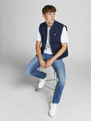 رجل يرتدي بنطلون جينز سكيني أزرق يجلس على كرسي، مرتديًا تيشيرت أبيض وسترة زرقاء بلا أكمام